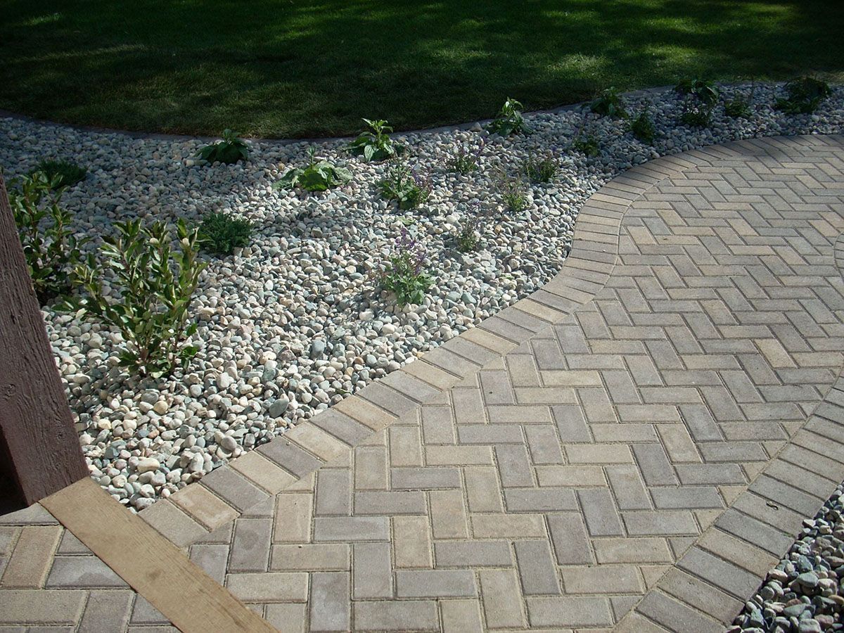 A herringbone-patterned paver walkway borders a gravel garden bed with small green plants