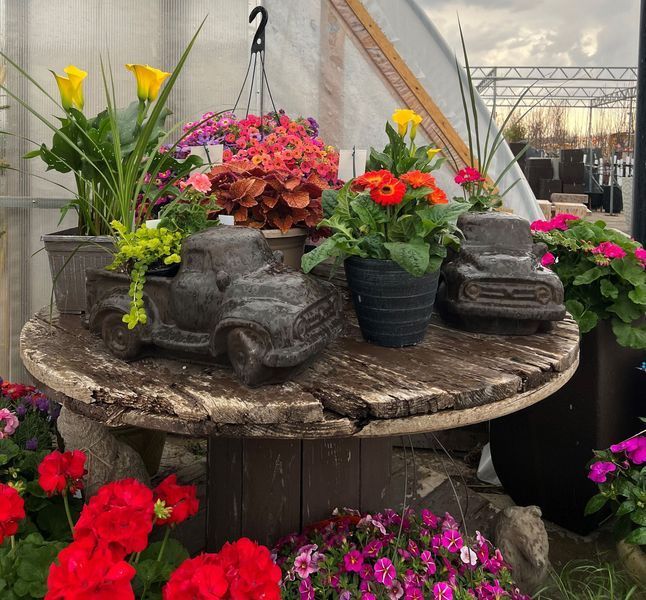 Flowers in planters and truck-shaped pots on a wooden spool table at a garden center.