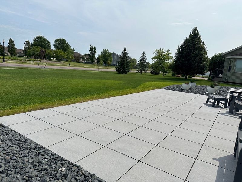 A paved patio with gray tiles, bordered by black rocks, overlooking a green lawn under a blue sky.