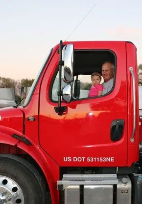 Man and child sit in red semi-truck cab, smiling.