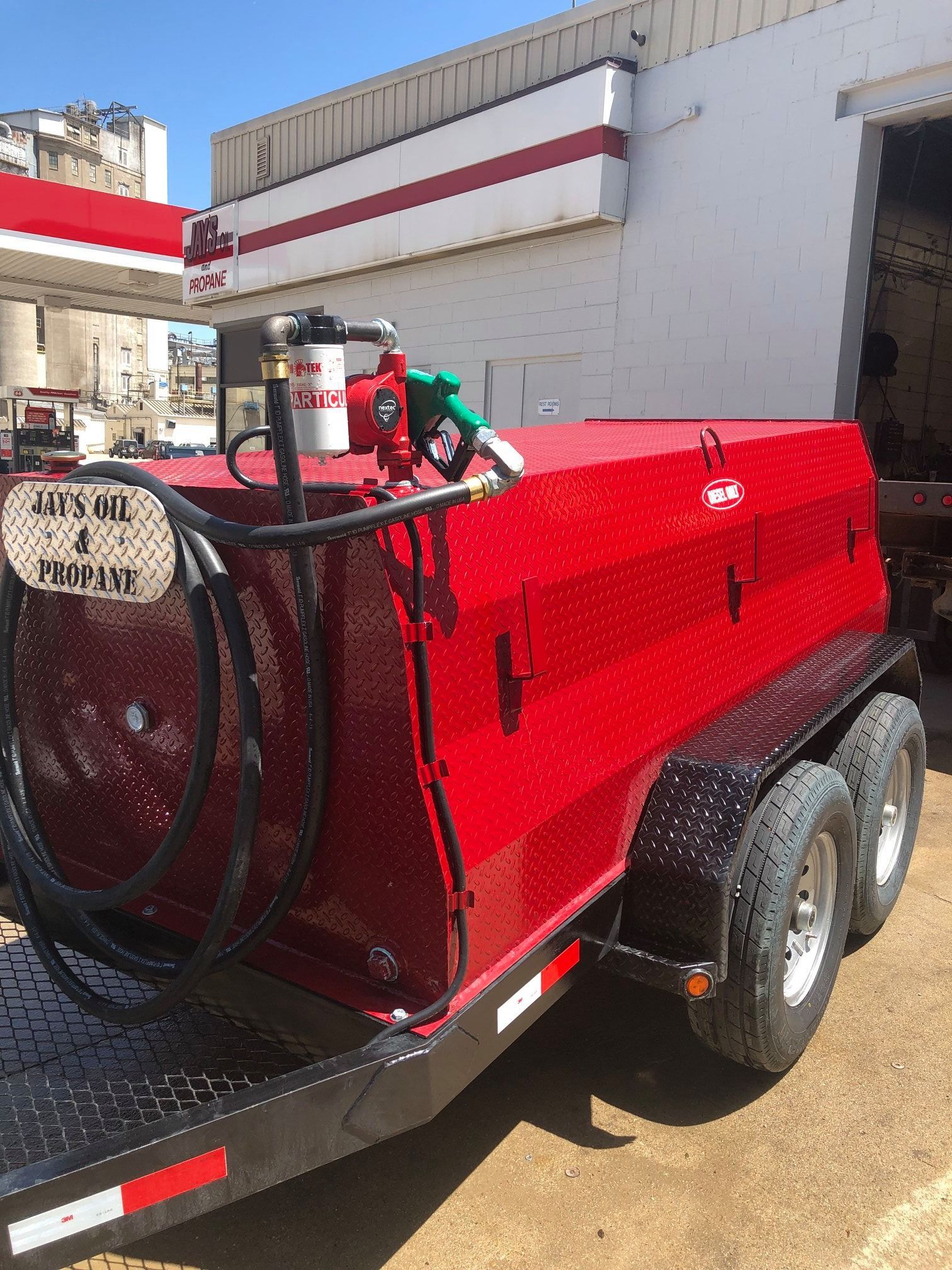 Red fuel tank trailer, with hose and nozzle, parked near a building and gas station.