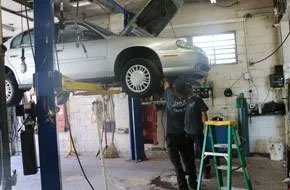 Silver car on a lift in a repair shop; two mechanics working underneath.