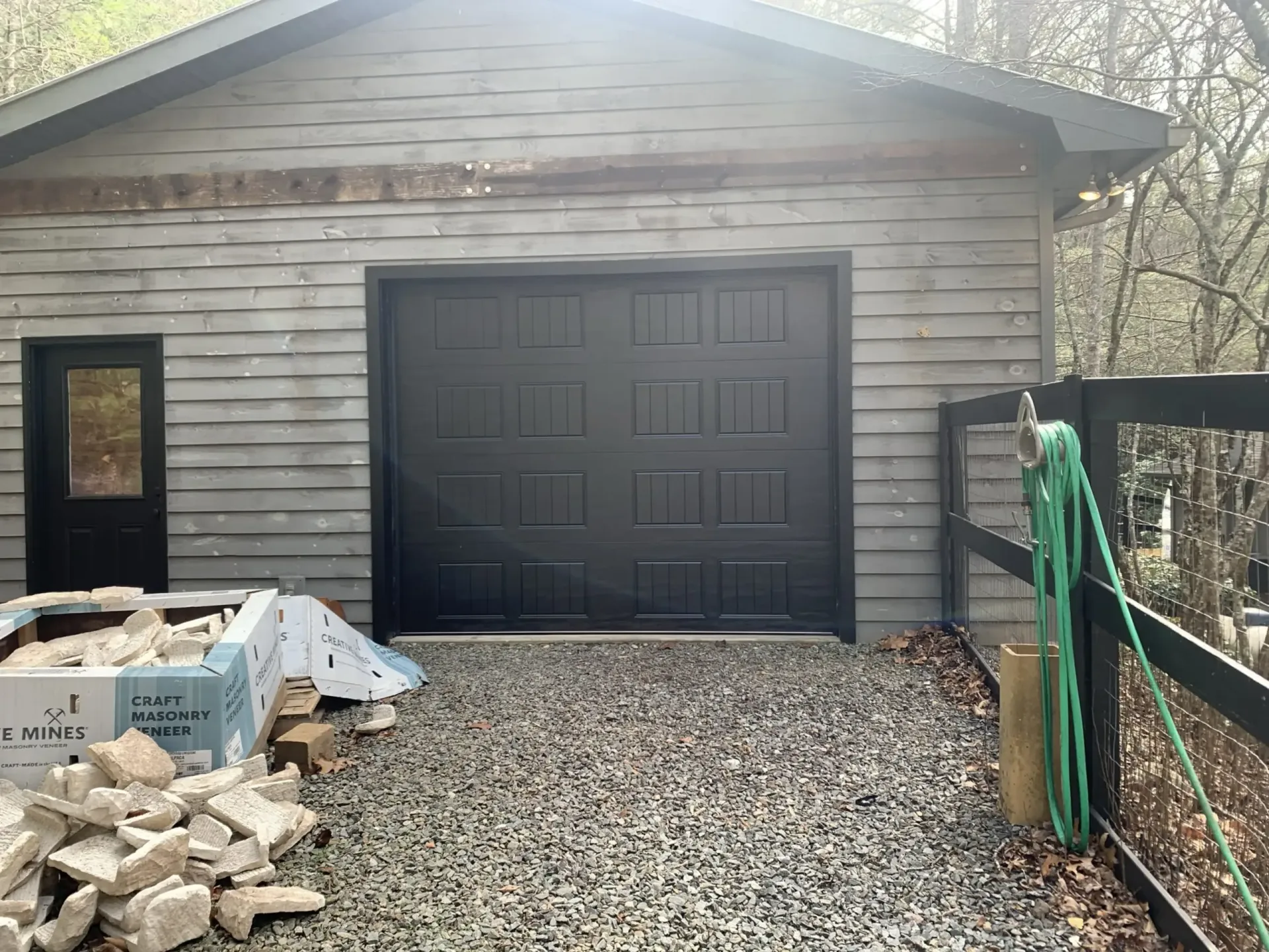 Detached wooden garage with a black door, gravel driveway, and stacked supplies beside it