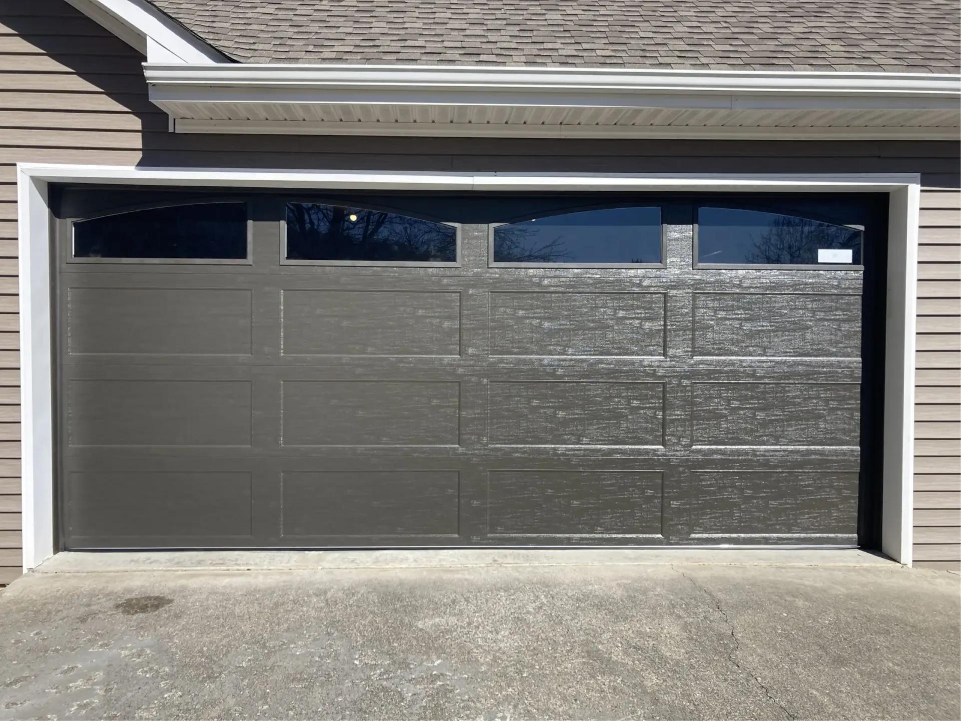 Dark gray garage door with four narrow windows, set in a beige house facade over a concrete driveway