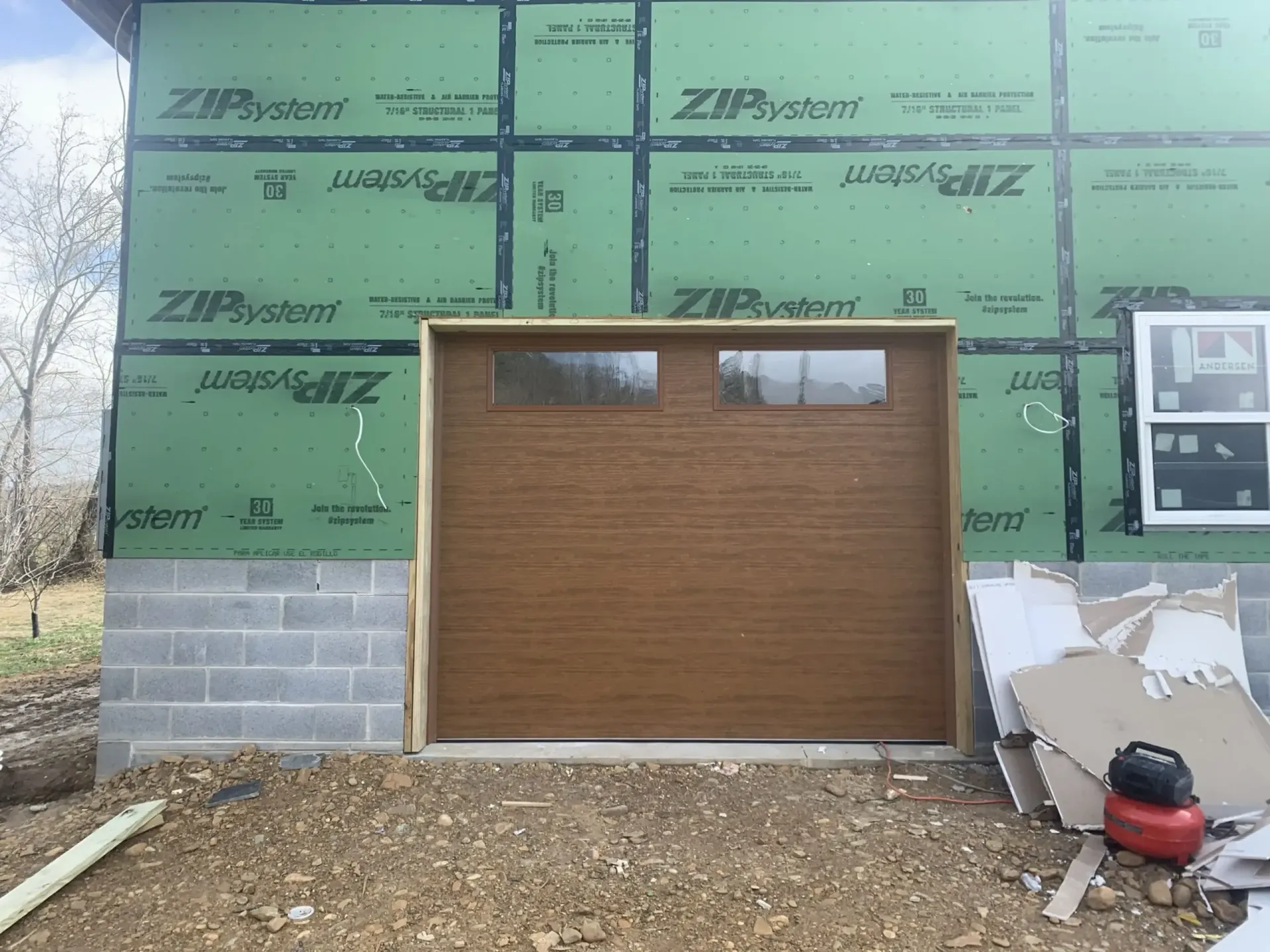 Brown garage door on a house under construction with green siding wrap and gravel driveway.