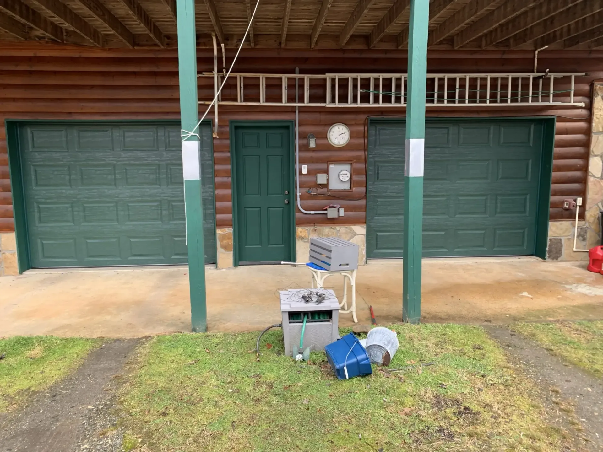 Green garage doors and front porch of a rustic cabin, with boxes and bags on the grass in front