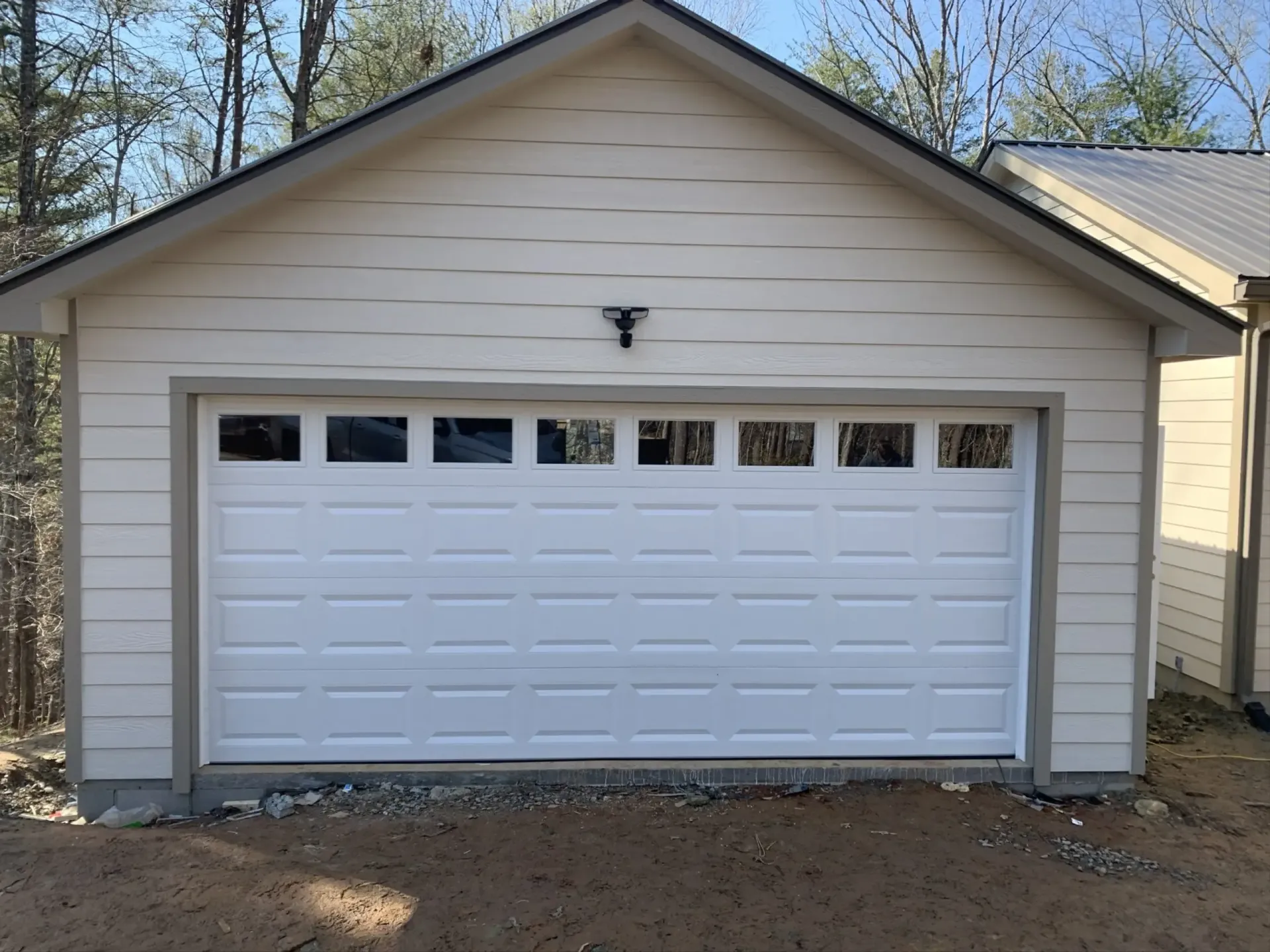 White garage with gabled roof and small upper windows, attached to a beige house in a wooded setting