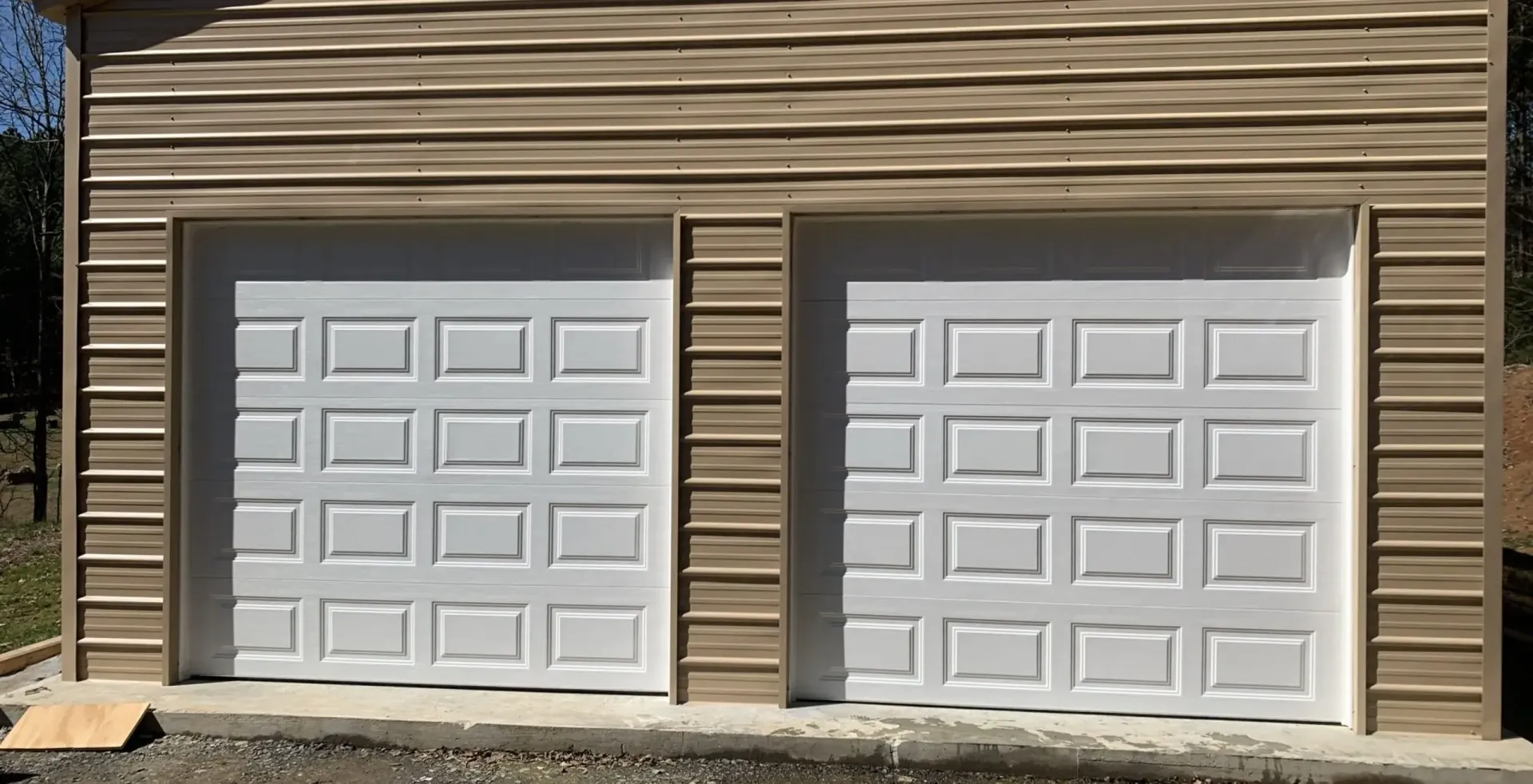 Two white garage doors on a tan-sided building with a paved driveway in front