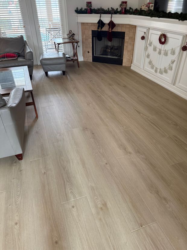 A light-toned wood-look floor in a living room with a gray armchair, small footstool, side table, and a corner fireplace.