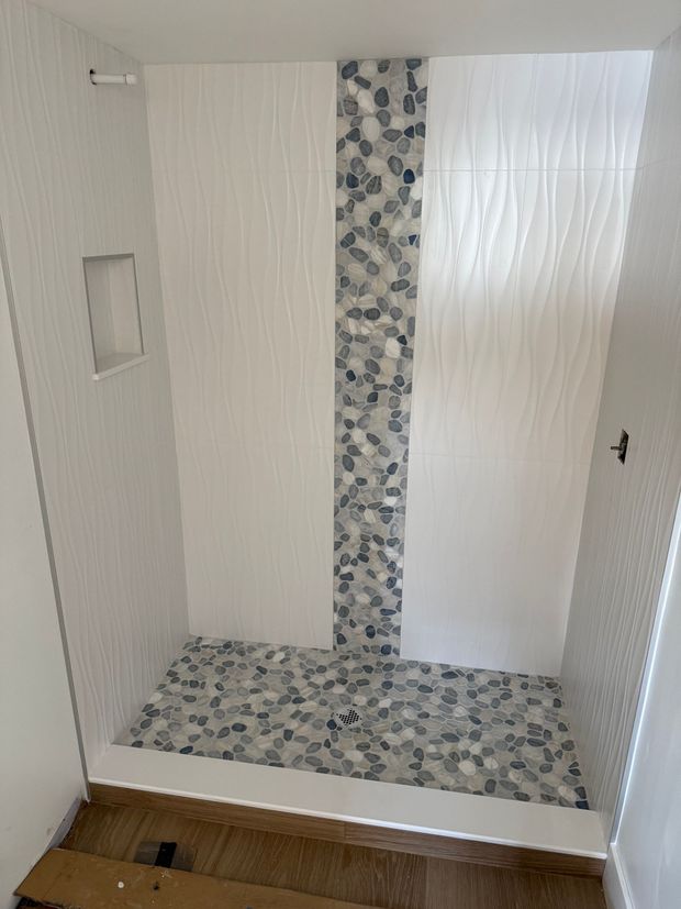 A modern shower stall featuring white textured wall tiles, a vertical gray pebble accent strip, and a matching pebble floor.