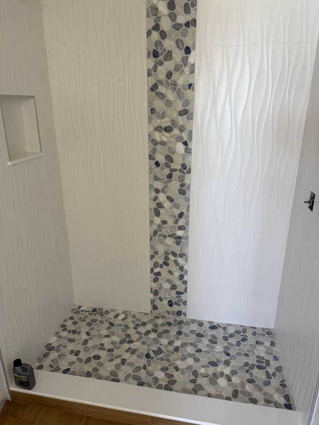 A shower stall with white, textured walls, a vertical strip of gray pebble tiles, and a matching pebble floor.