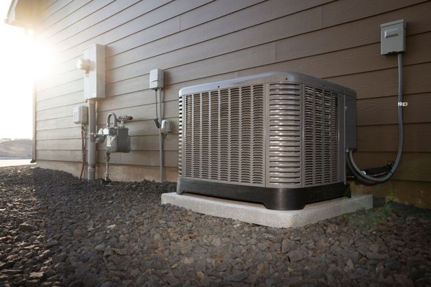 Outdoor HVAC unit beside a house wall, with sunlight and gravel ground.