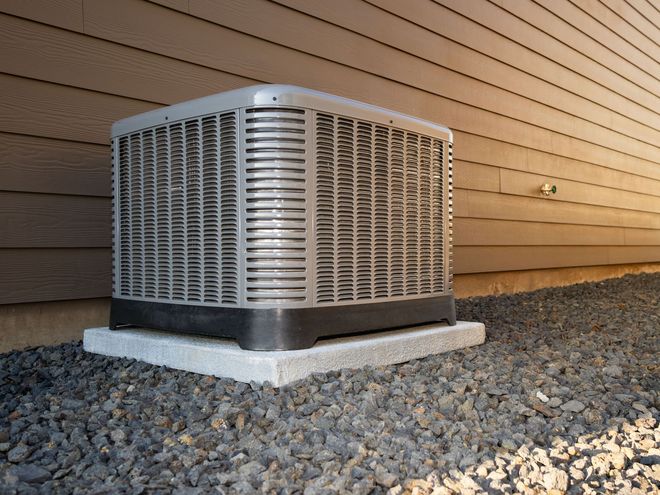 Outdoor air conditioner unit beside a house wall on gravel, with brown siding and white stone base.