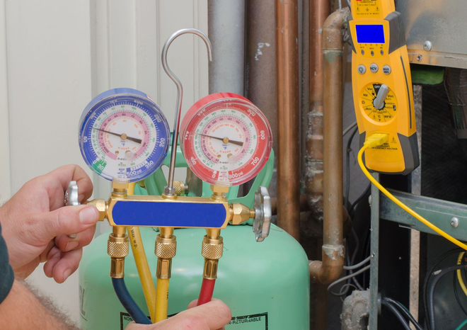 Hand holding HVAC manifold gauges with blue and red dials beside a yellow digital meter and pipes.