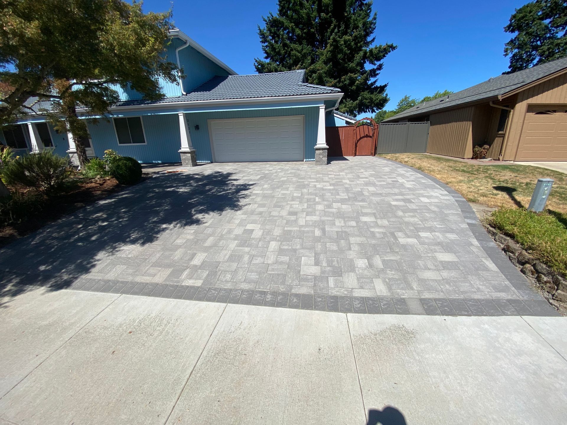 Paver driveway leading to a blue house with a two-car garage. A sidewalk borders the driveway.