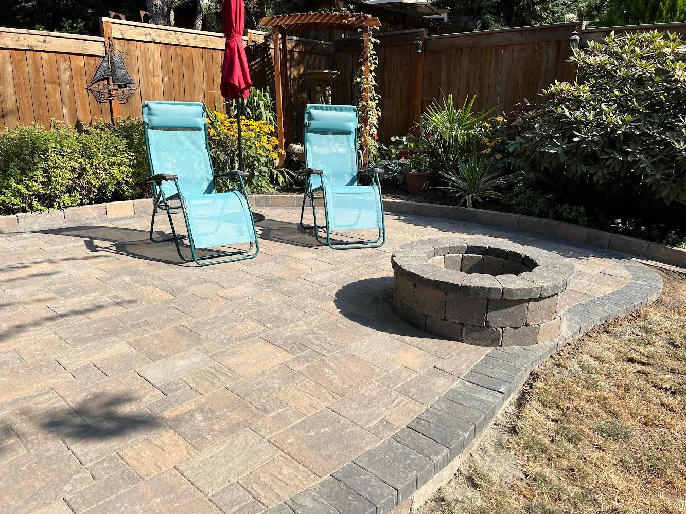 Paver patio with fire pit, two blue chairs, and a red umbrella in a backyard.