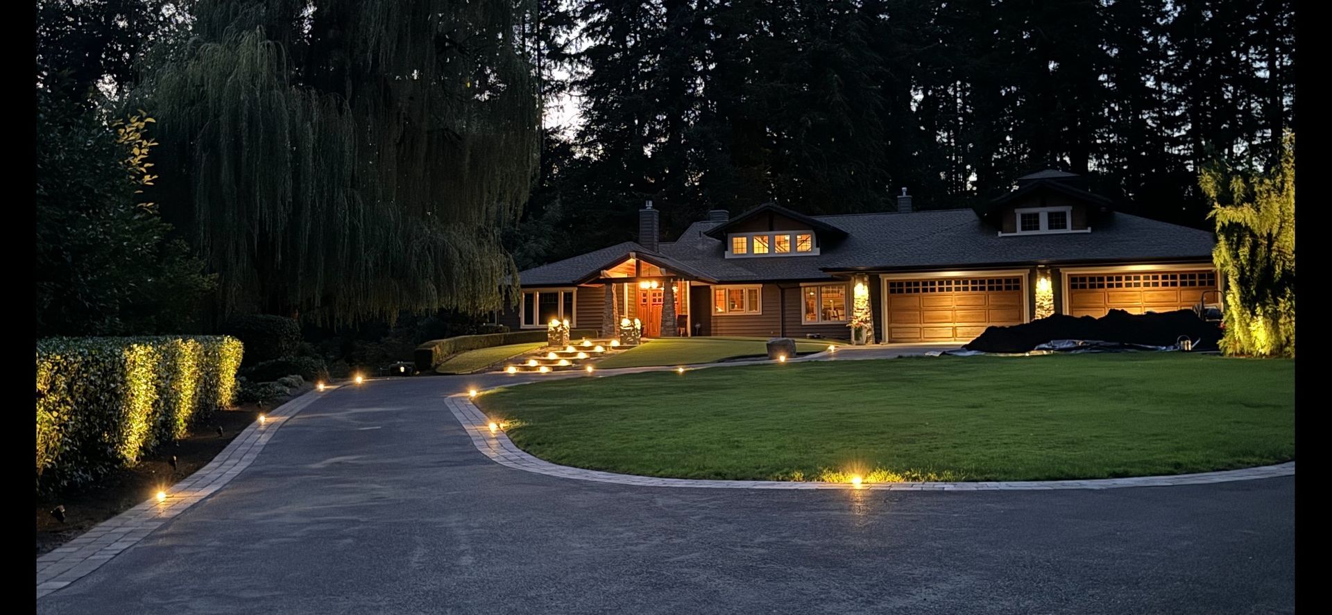 House exterior at night, driveway with lights, lawn and surrounding trees.
