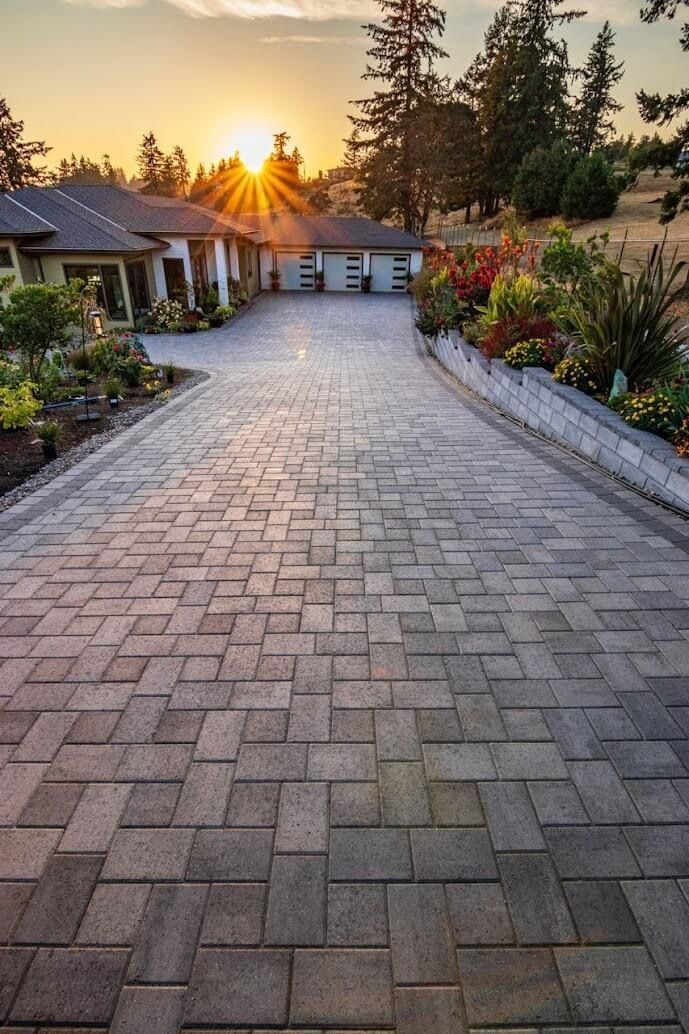 Brick driveway leading to a house with garage, flowers, and sunset in the background.