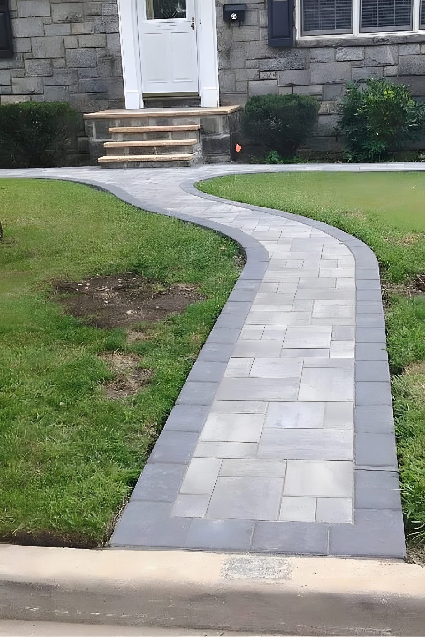 A curved stone walkway leads to the entrance of a house with stone siding. The path has a gray border and green grass on either side.