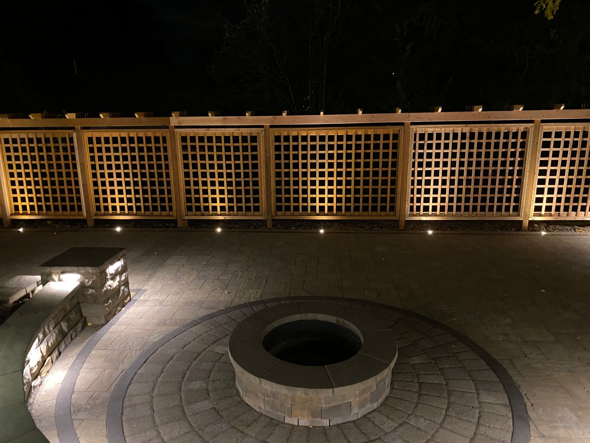 Outdoor patio at night, with a fire pit, illuminated fence, and stone paving.