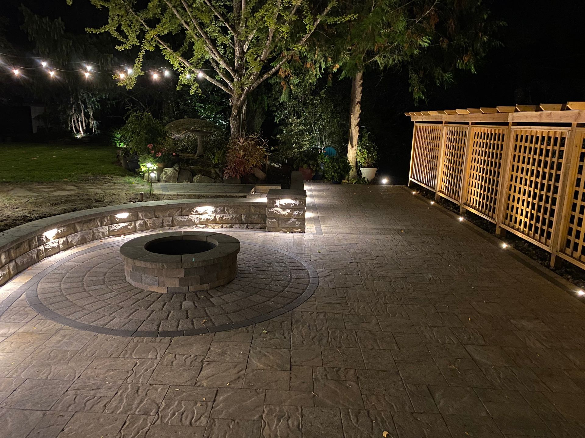 Nighttime outdoor patio with fire pit, lighting, and wooden trellis.