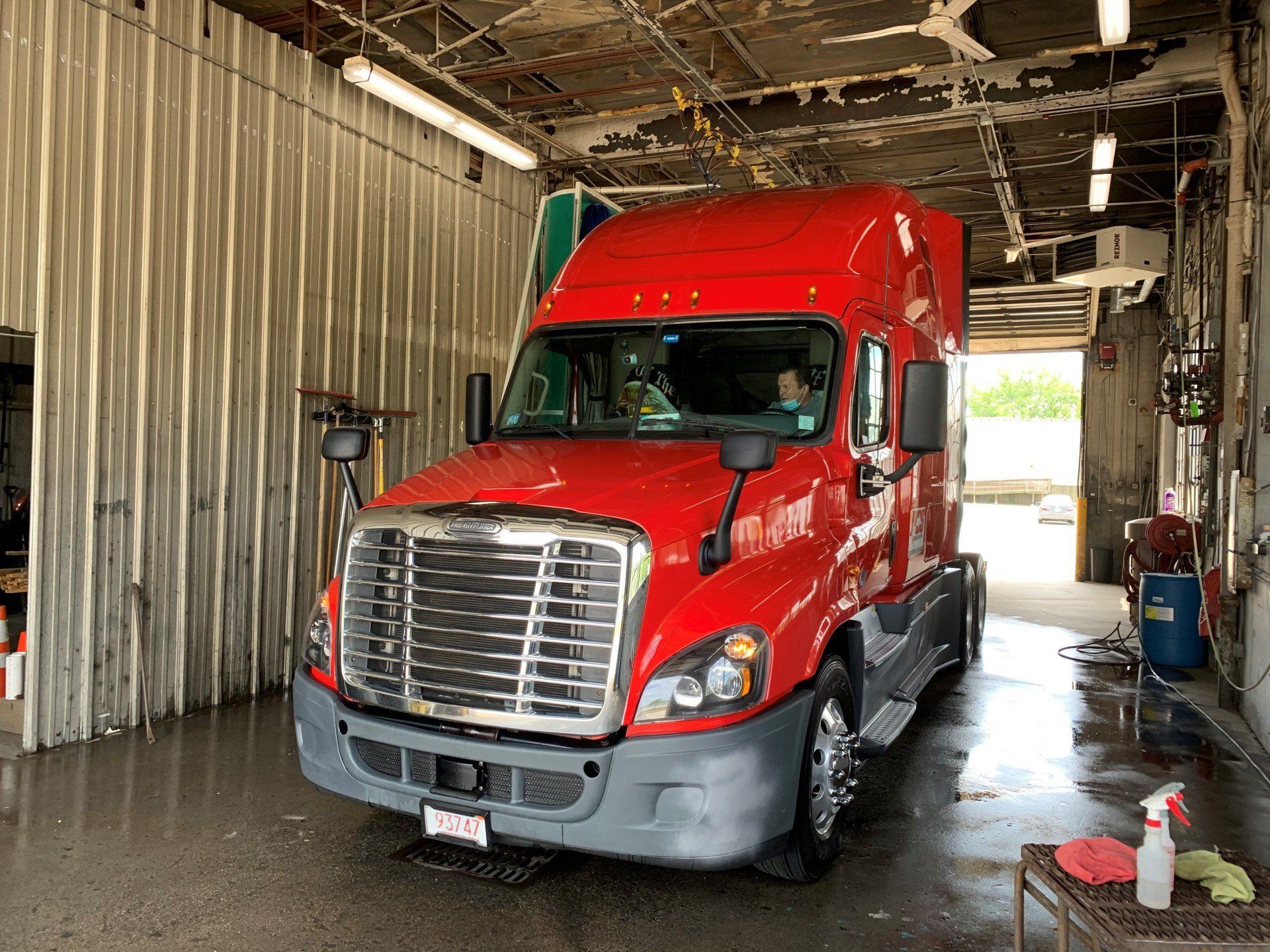 Red semi-truck parked inside an industrial garage with corrugated metal walls and wet floor.