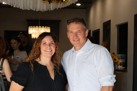 Two people smiling indoors at a social gathering, with chandeliers and dark walls in the background.