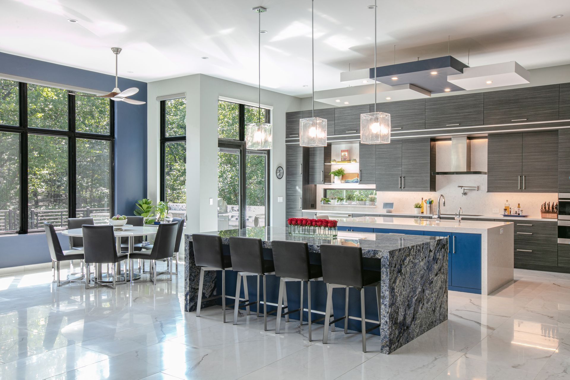 Modern kitchen with blue and gray cabinetry, marble island and floors, dining area with large windows.