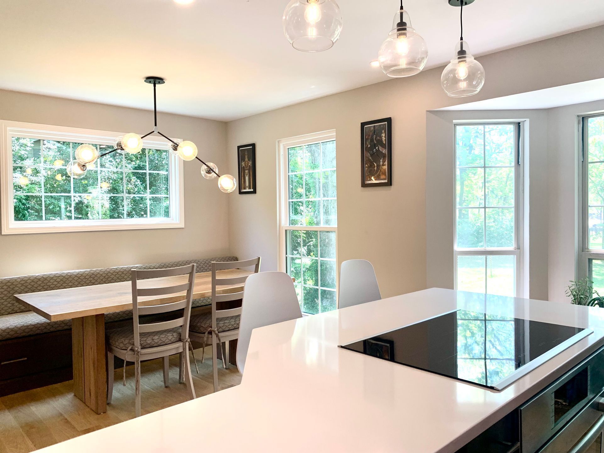 Modern kitchen with island, dining nook, and windows. Light fixtures hang above.