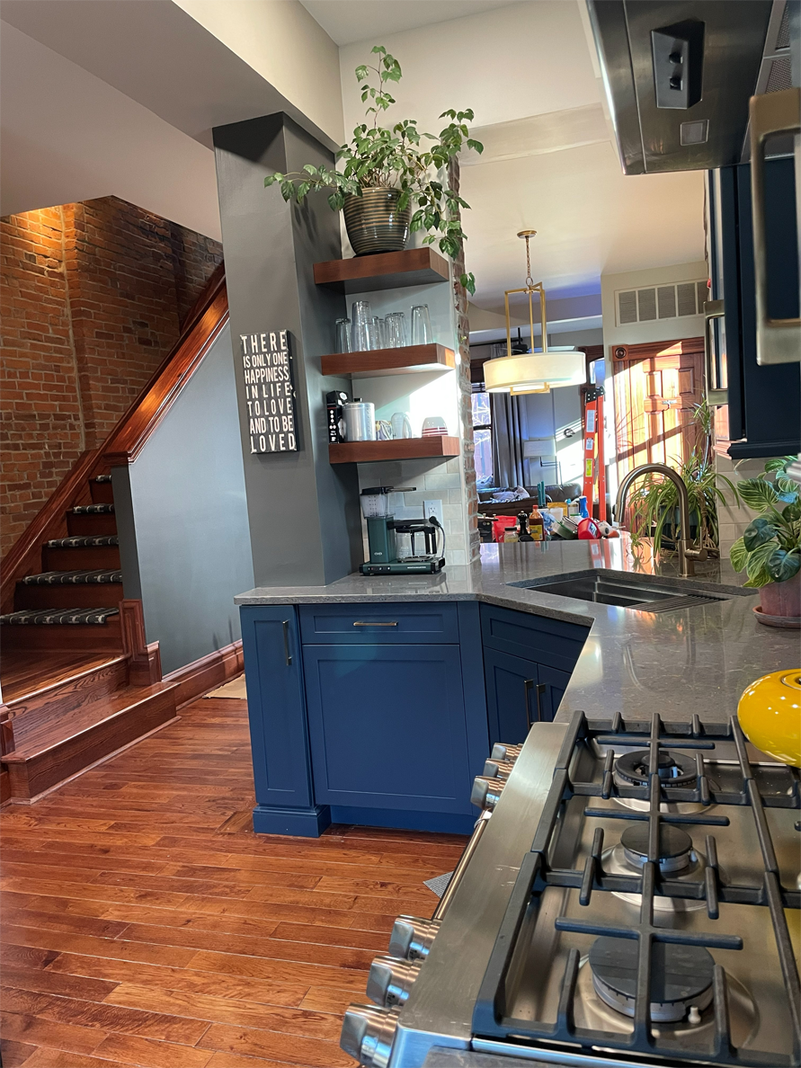 Kitchen interior with wooden floors, brick staircase, and blue cabinets.  Floating shelves hold glasses and plants.