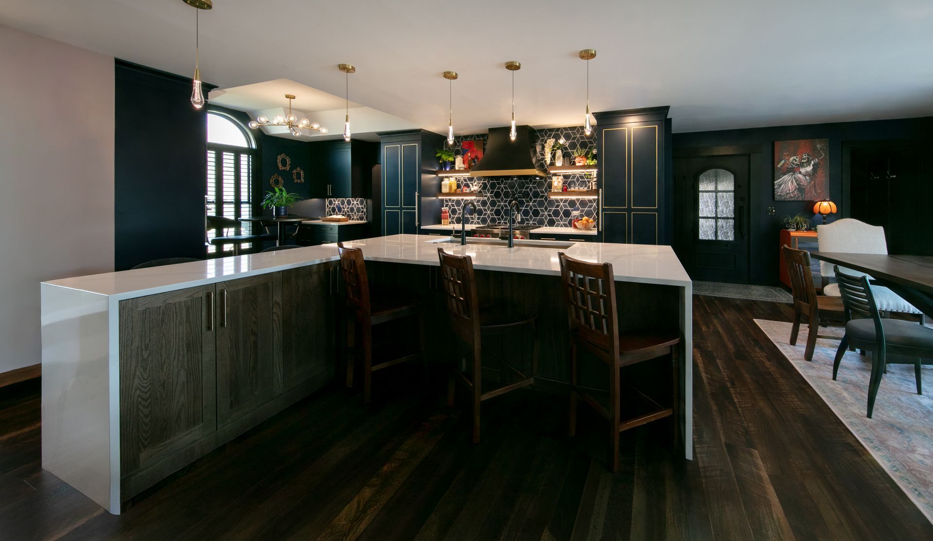 Dark kitchen with island, wooden floors, and hanging lights.  Cabinets and accents in dark blue.  Dining table in foreground.
