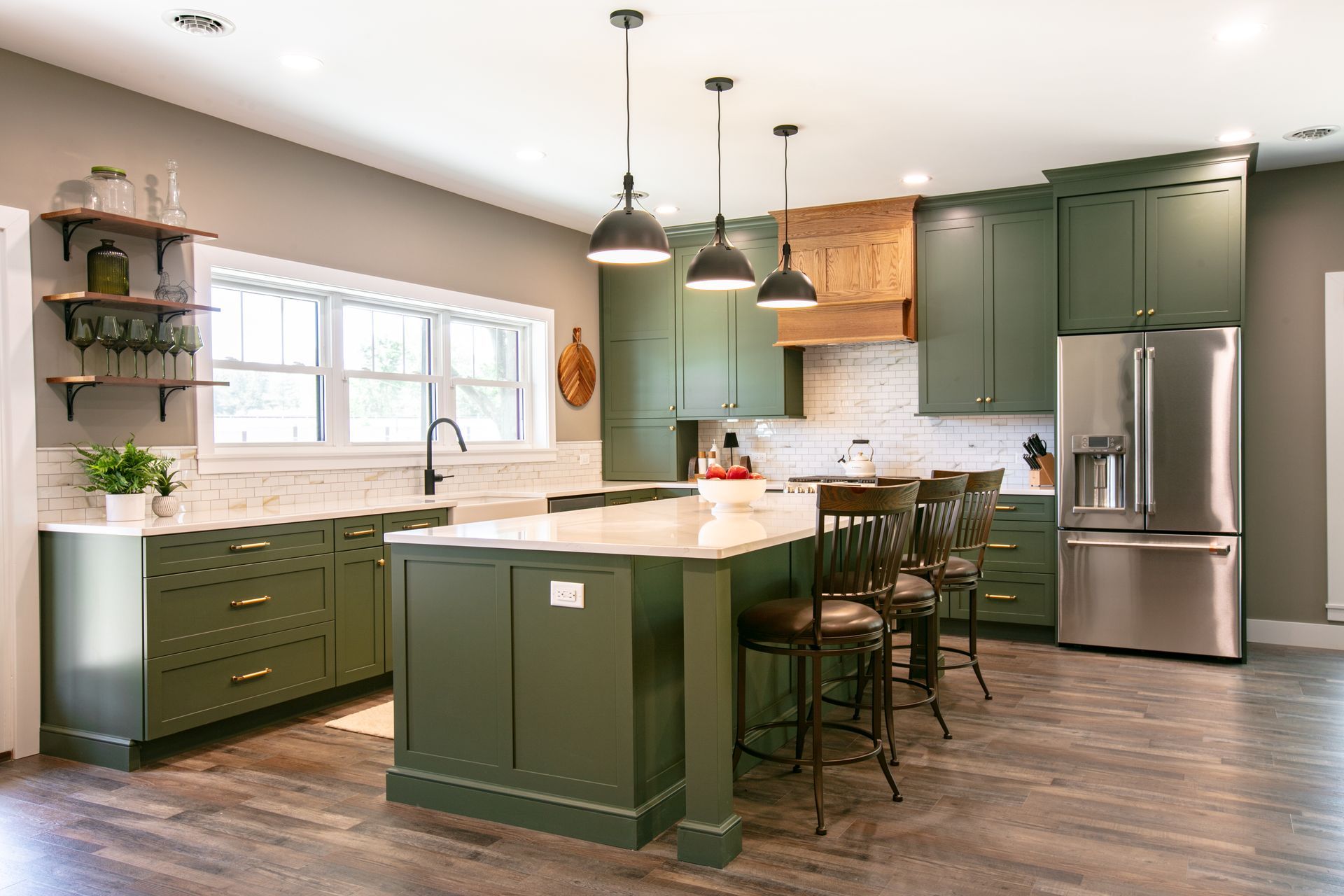 Green kitchen with island and stainless steel refrigerator. Three pendant lights hang above.