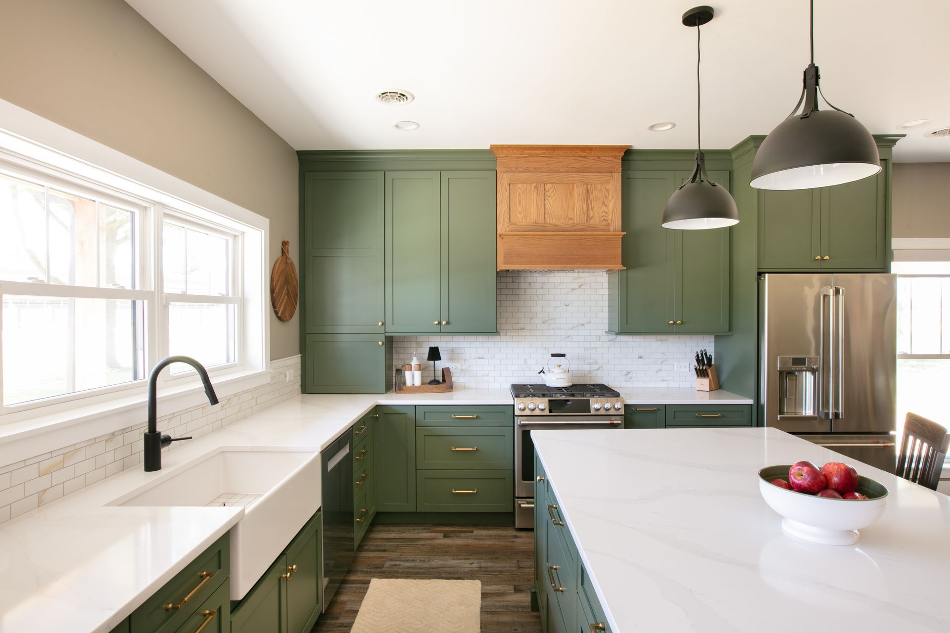 Green kitchen with white countertops, wooden range hood, and stainless steel appliances.