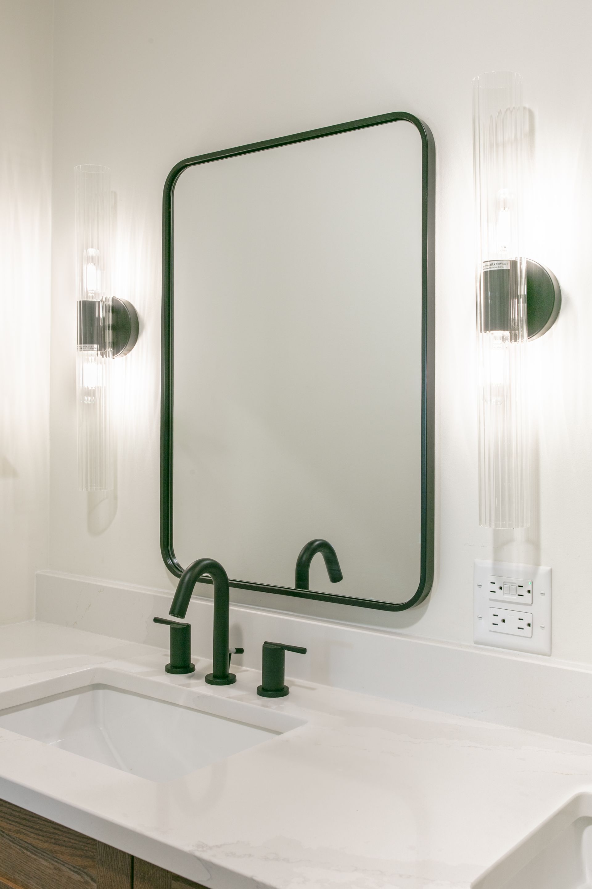 Bathroom vanity with a black-framed mirror, faucet, and sconces, white countertop, and sink.