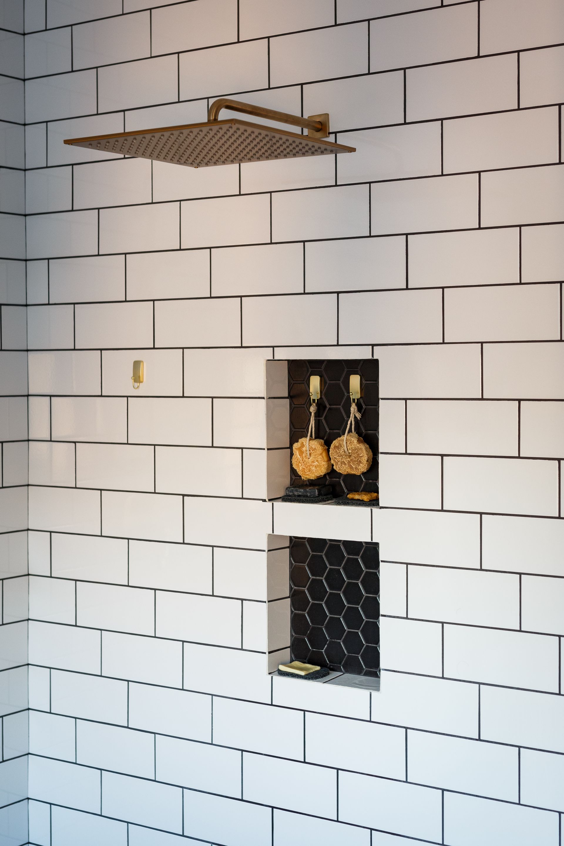 White tiled shower with brass fixtures, including a square showerhead. Two recessed shelves hold soap and shower supplies.