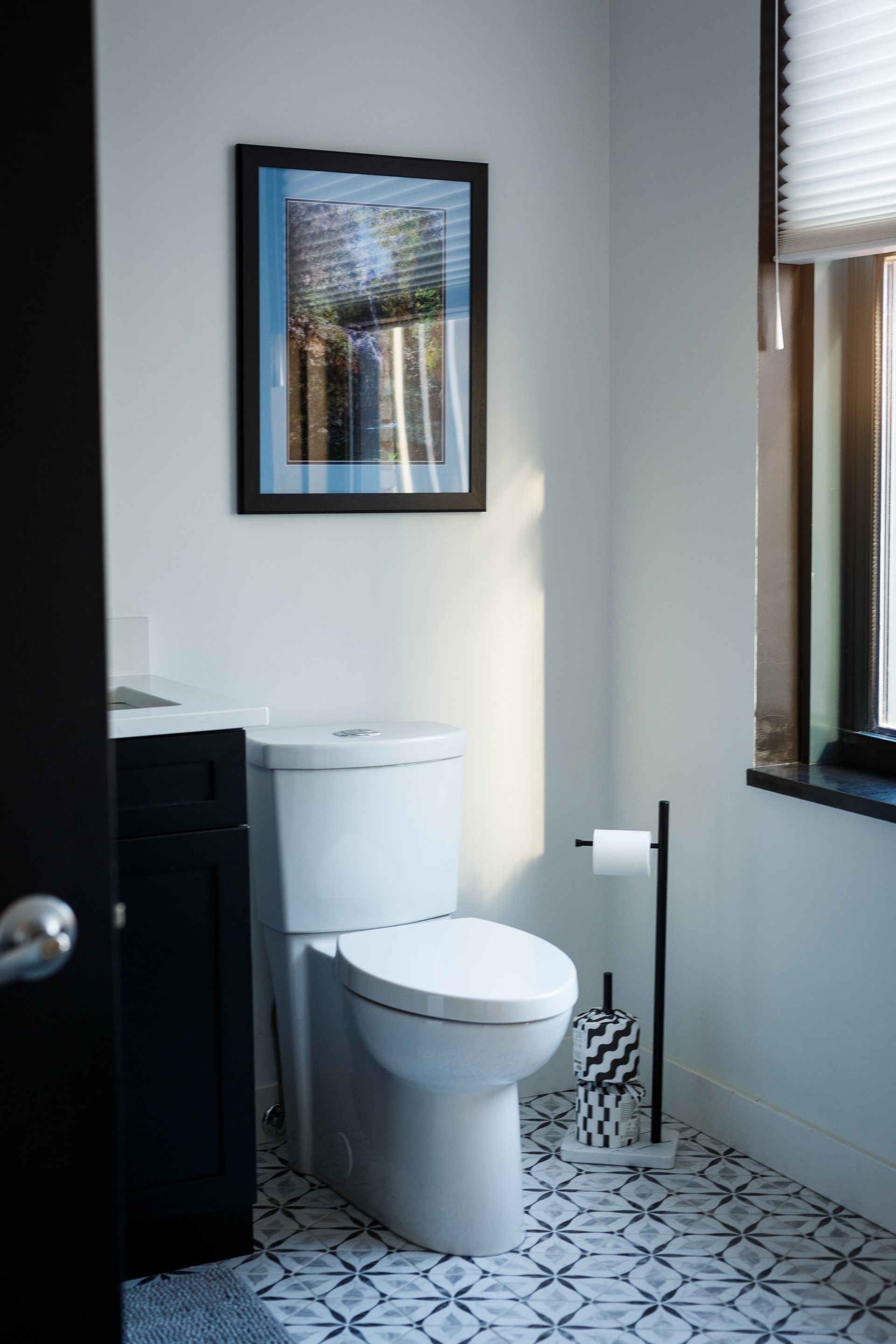 Bathroom with a white toilet, art, and patterned floor tiles. Sunlight through the window.