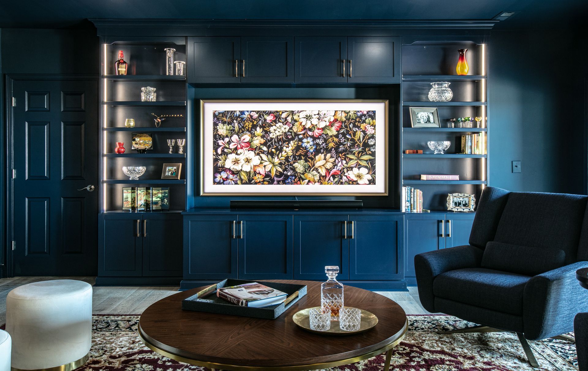Dark blue built-in media center with TV, shelves, and cabinets. A dark armchair and round coffee table in front of the unit.