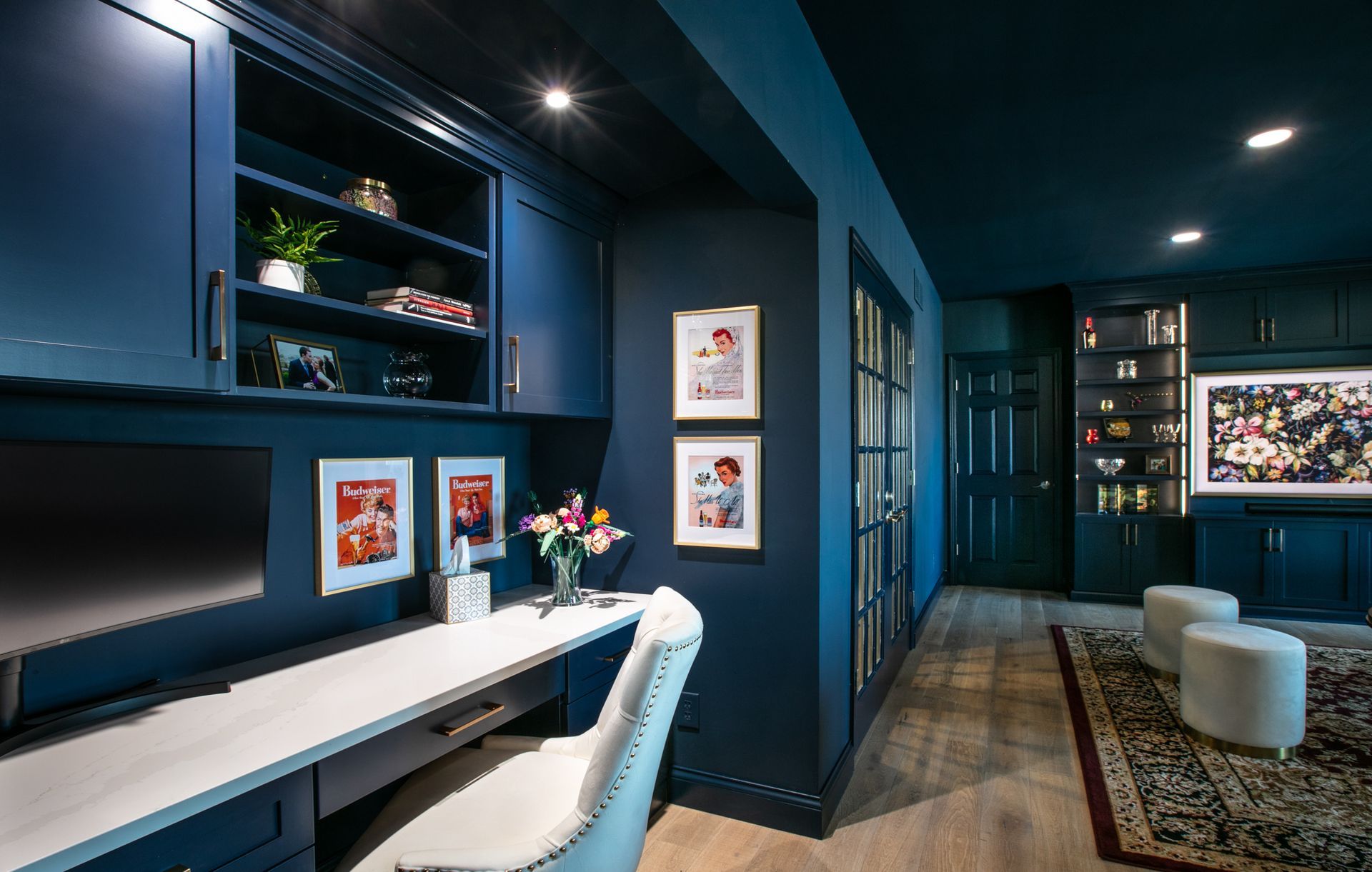 Dark blue home office with built-in desk, cabinets, and a rug. Features desk chair, artwork, and recessed lighting.