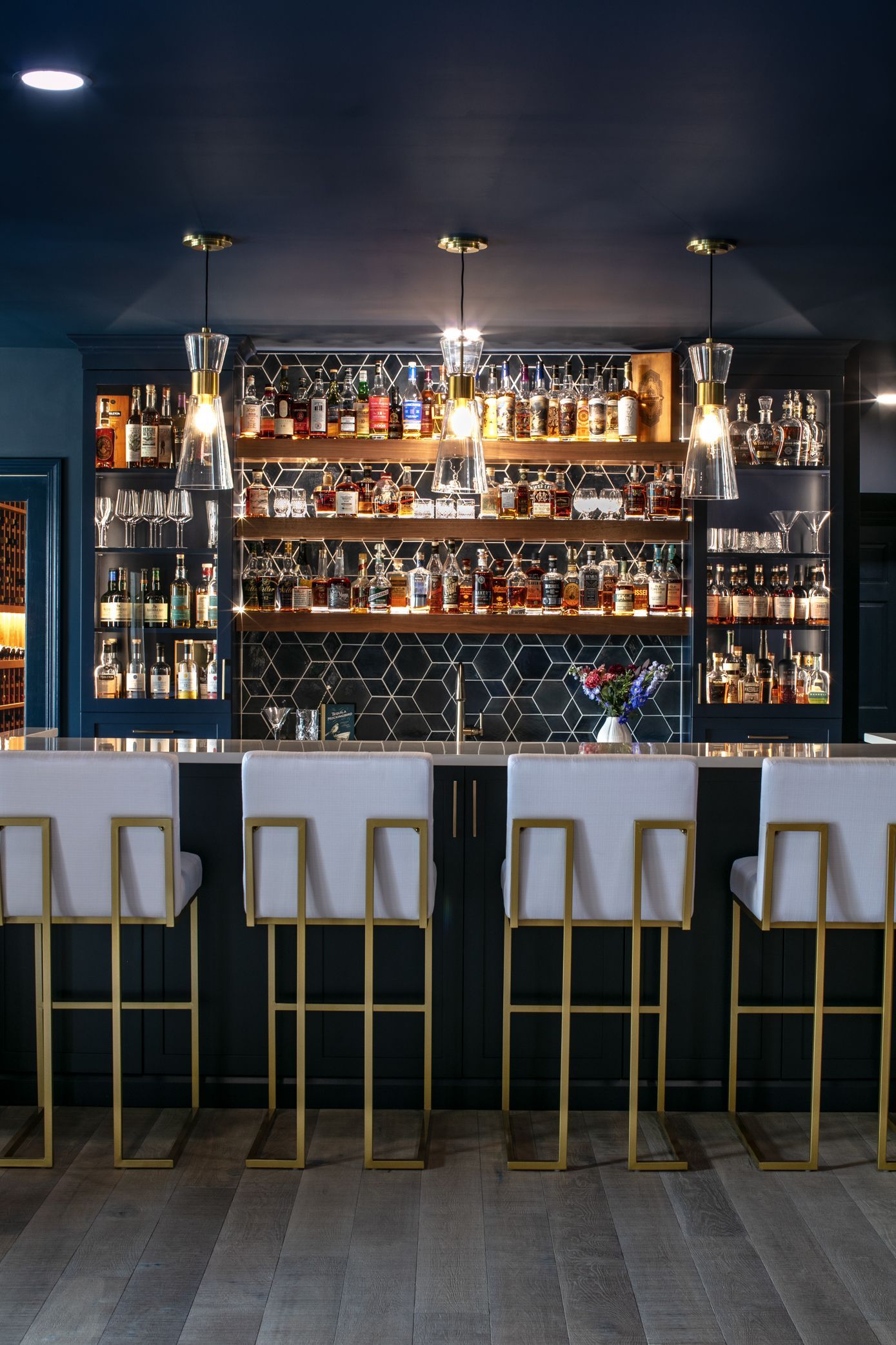 Bar with backlit liquor shelves, white and gold bar stools, dark blue ceiling, and wooden floor.