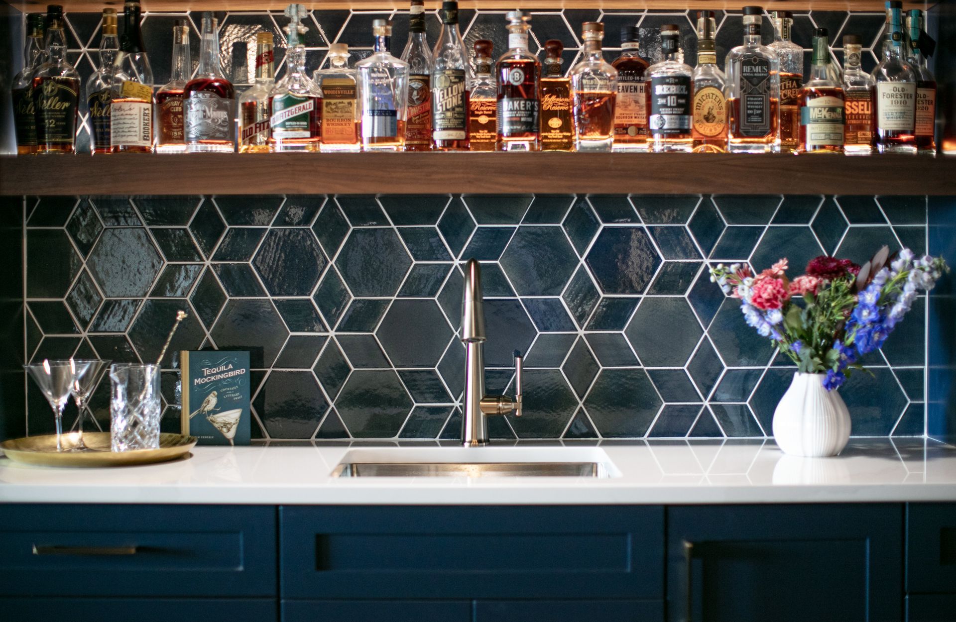 Bar with blue cabinets, hexagonal tile backsplash, liquor bottles, sink, faucet, and flowers.