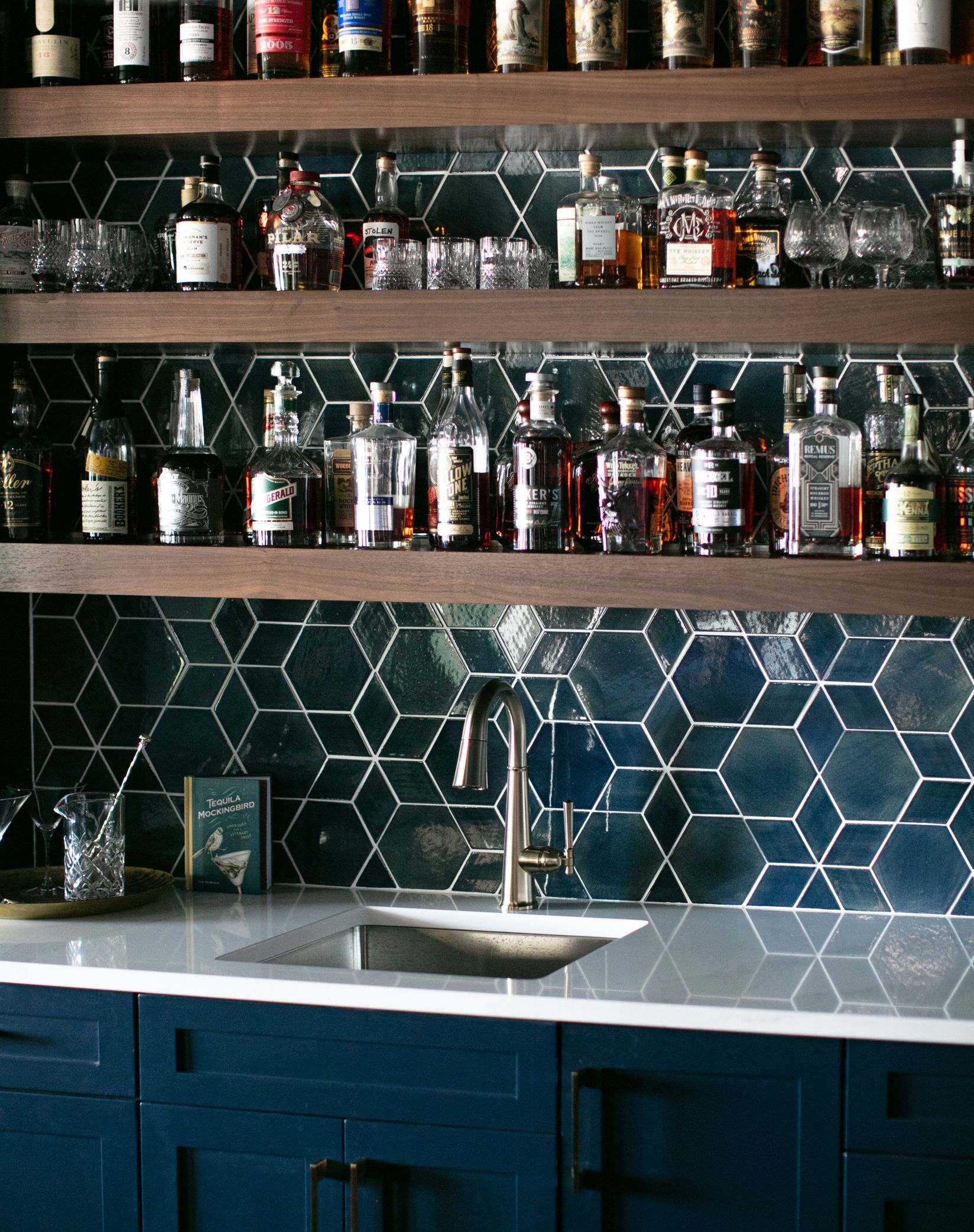 Bar area with blue hexagonal tile backsplash, wooden shelves stocked with liquor bottles, and a stainless steel sink.