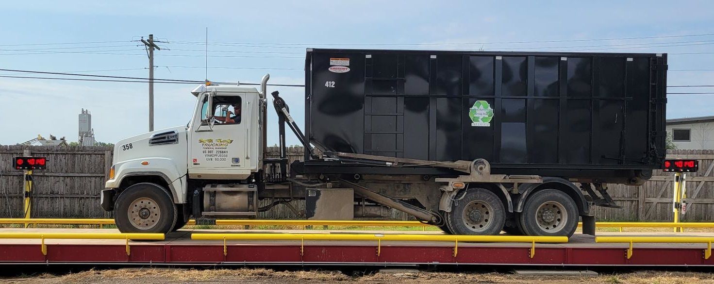 White dump truck hauling a black container on a flatbed trailer beside train tracks.