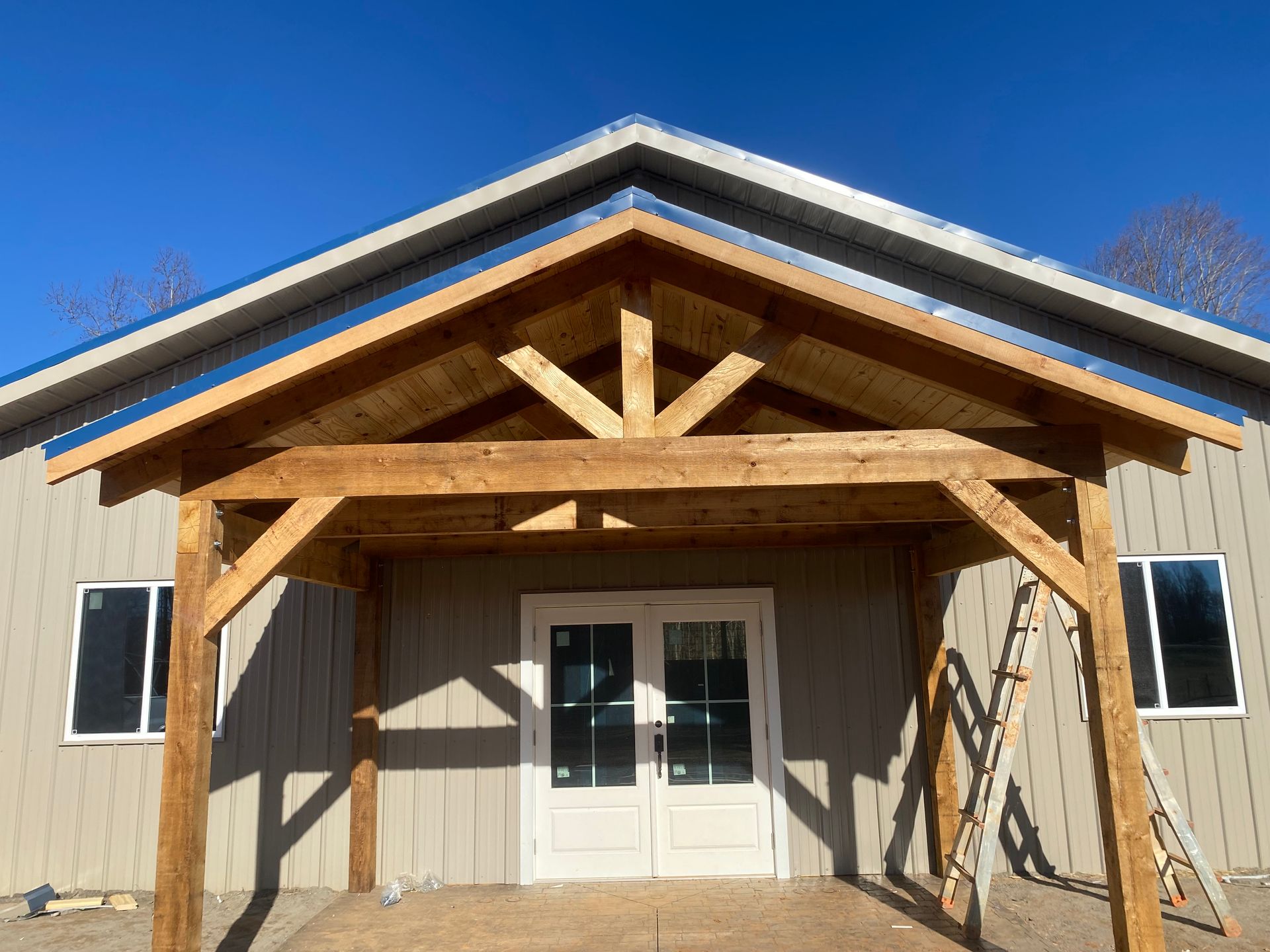 A wooden porch shelters double doors of a beige building with a blue metal roof, under a clear blue sky.