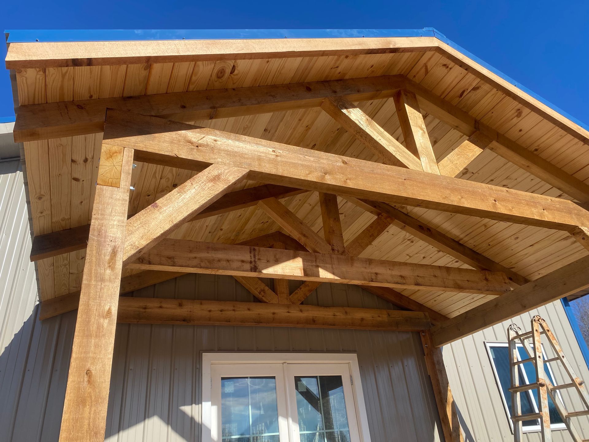Wooden porch with a gabled roof over a white door and windows. Light wood beams and rafters support the roof.