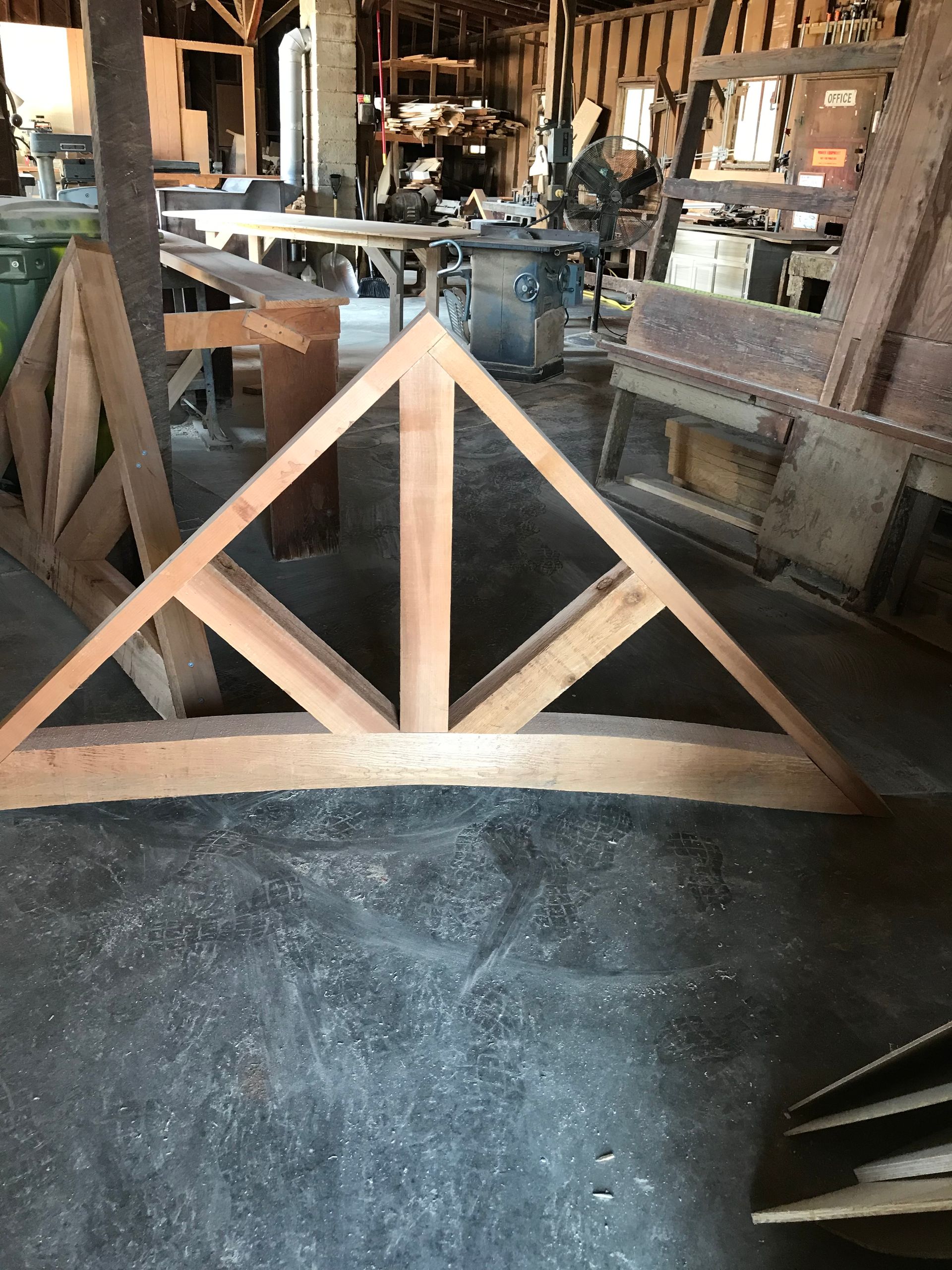 Wooden triangular truss on a workshop floor. Sawdust and other tools are visible in the background.