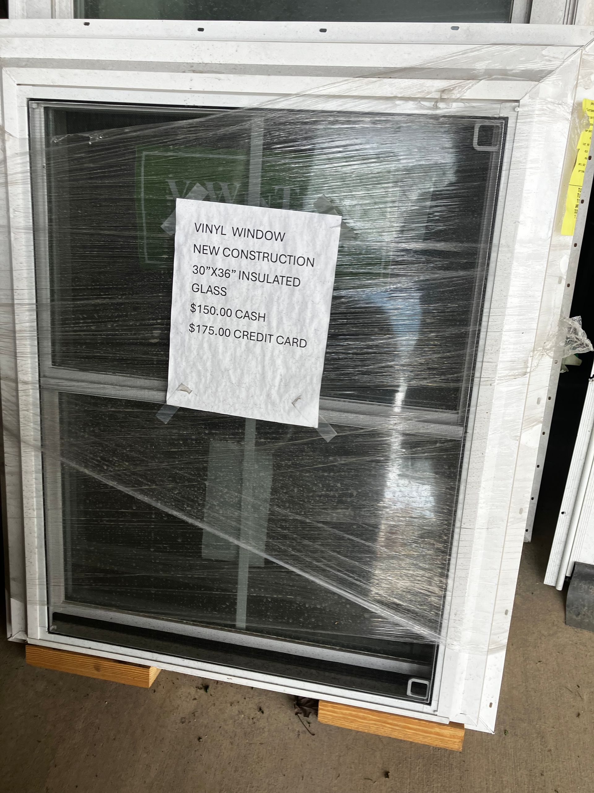 White-framed window wrapped in plastic with a white paper attached, likely containing details. It sits on a wooden pallet.