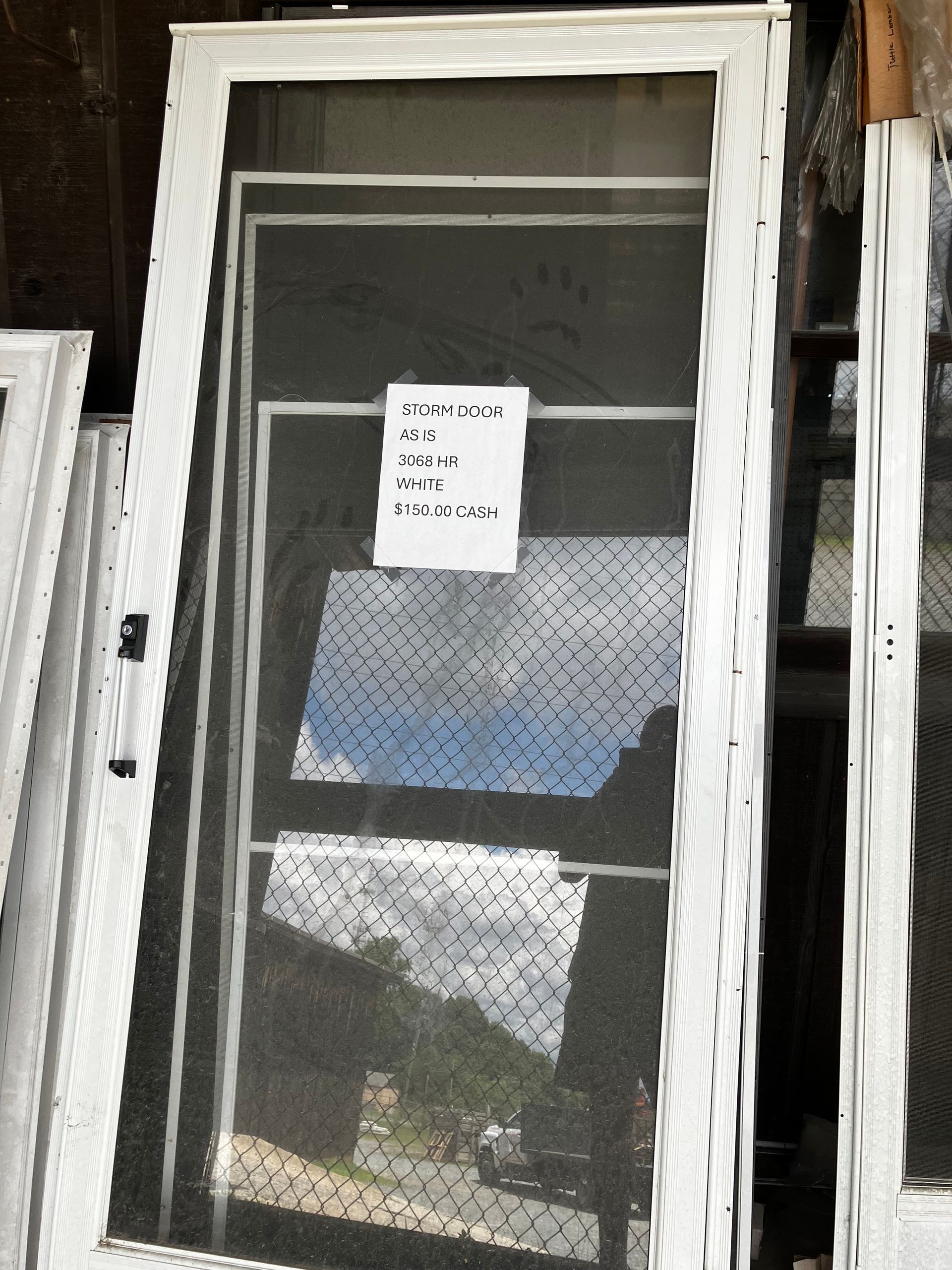 White-framed screen door with a note on it, leaning against other doors. The screen reflects a partly cloudy sky and a hillside.