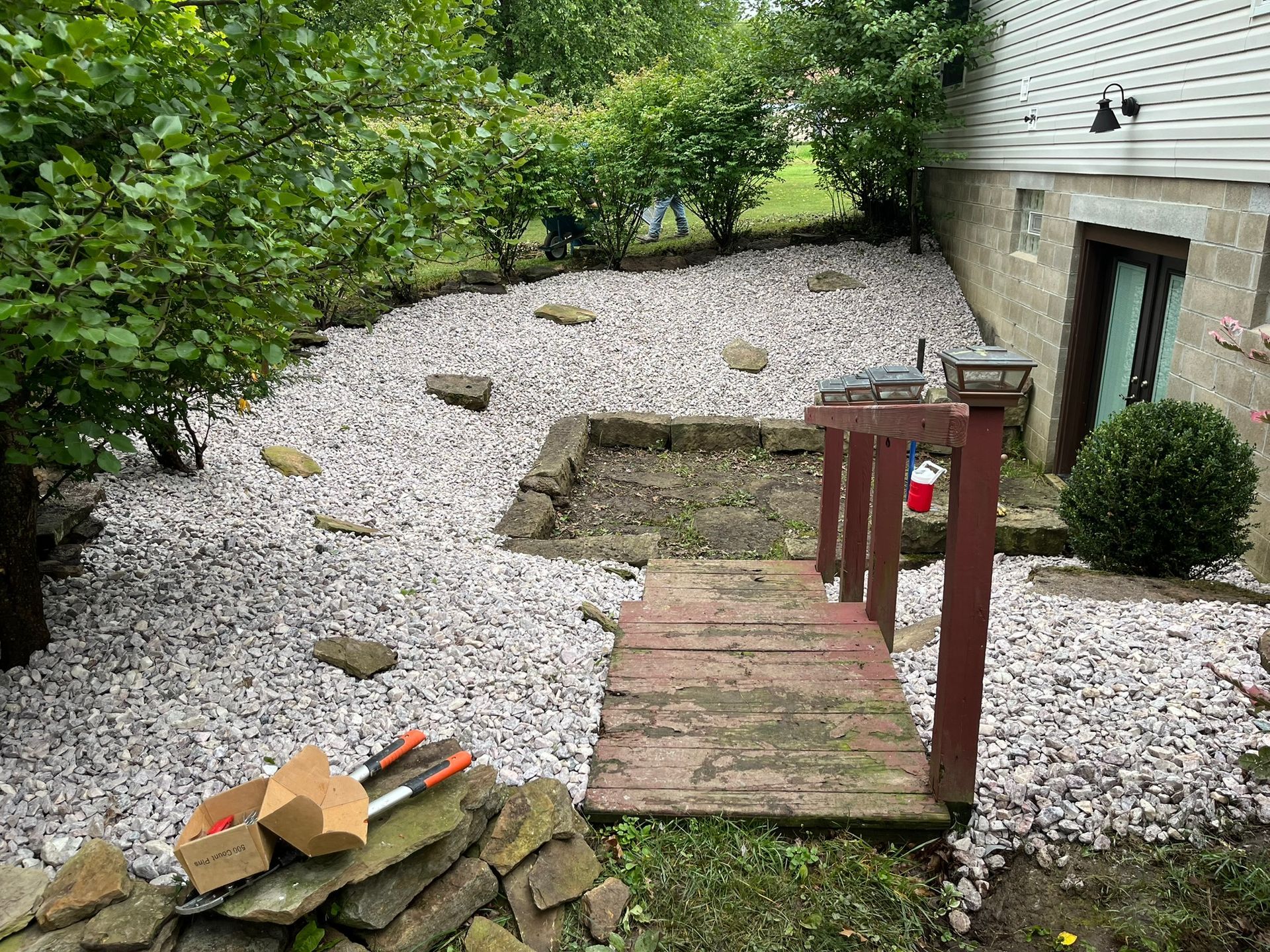 A wooden bridge leads to a gray gravel yard, with a building on the right and shrubs along the back.