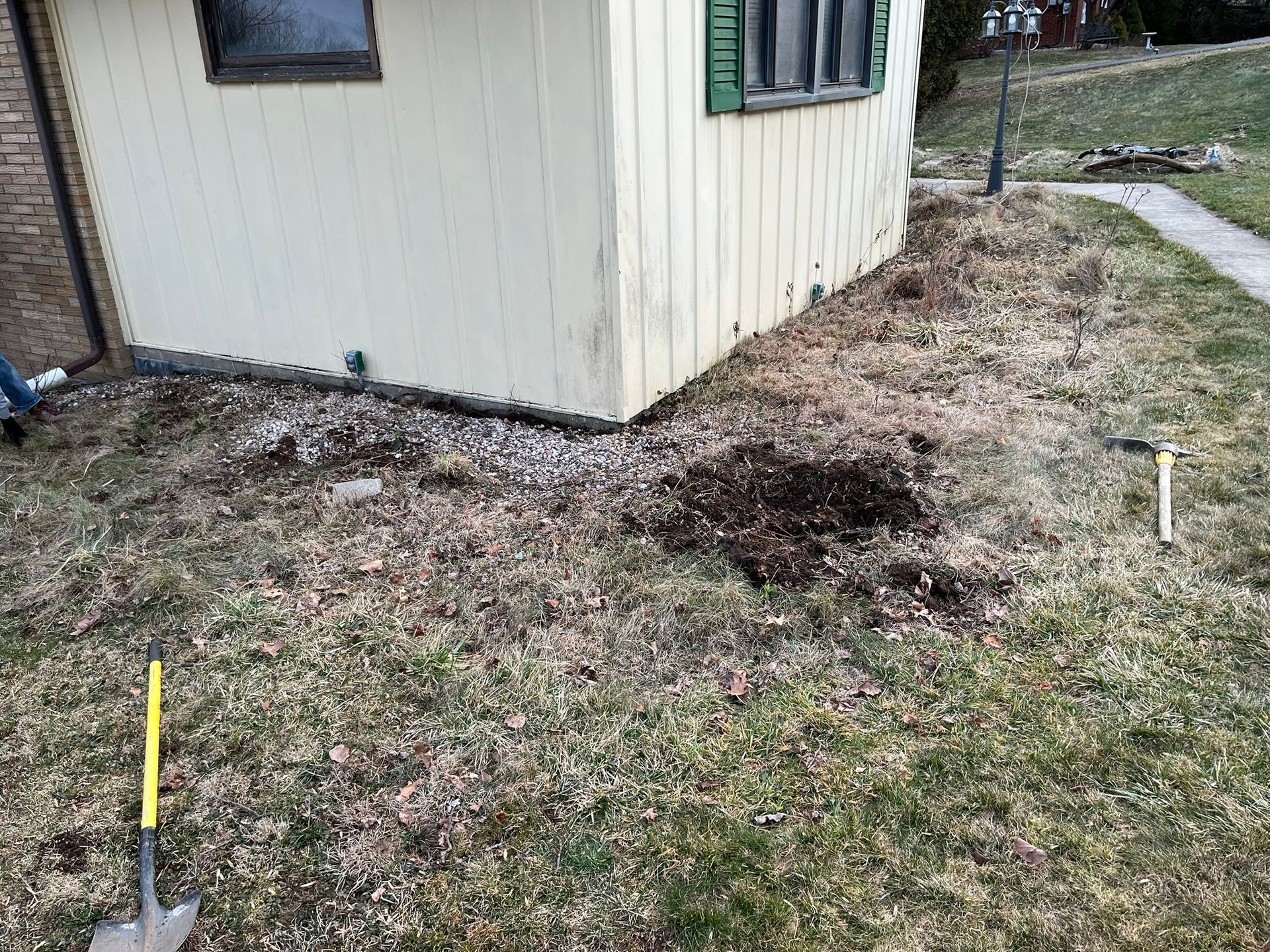 A building's corner next to patchy grass and a small garden bed with a shovel.
