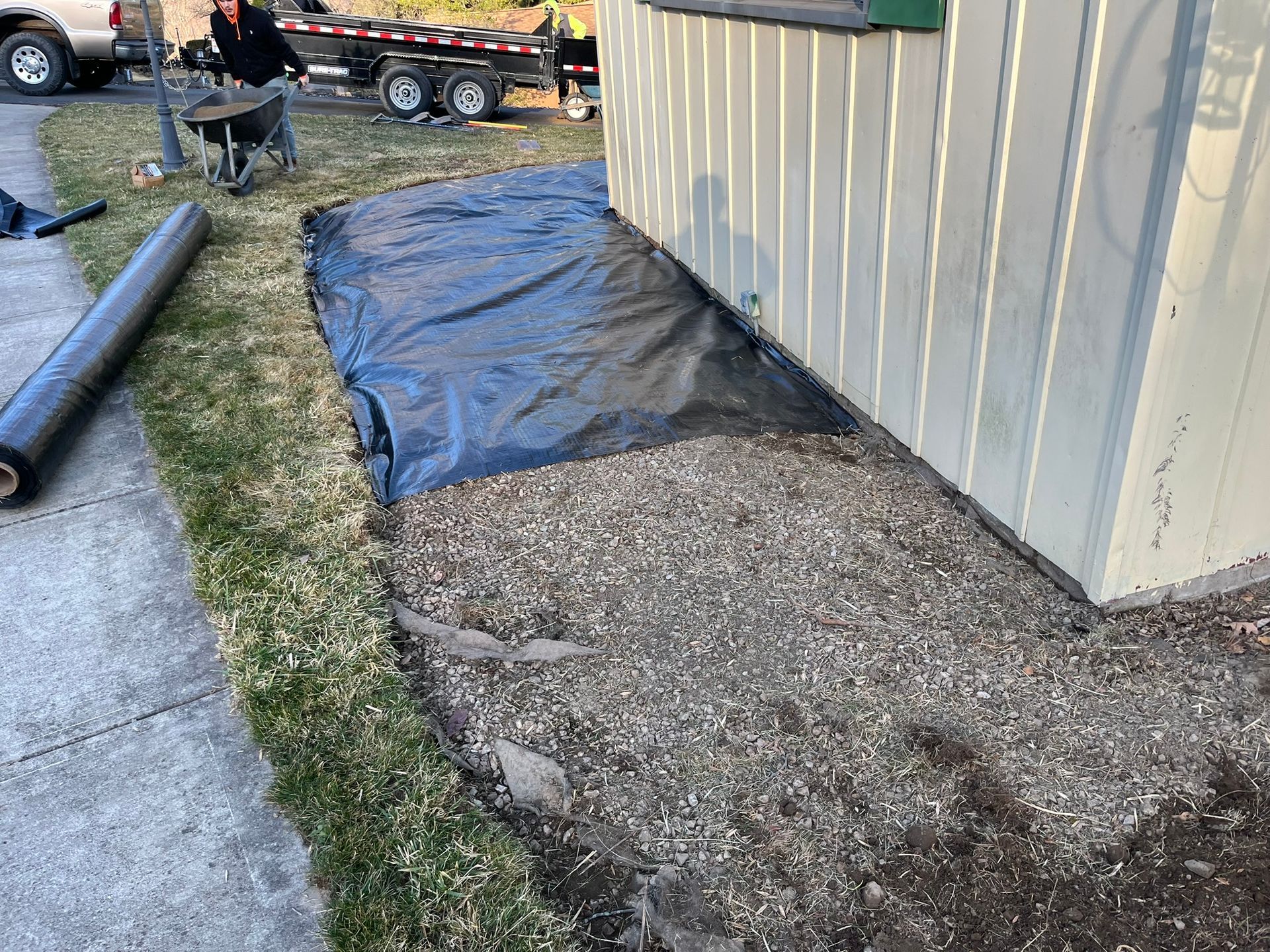 Gravel bed beside a building with black fabric underlay, fresh lawn edge, roll of fabric on grass.